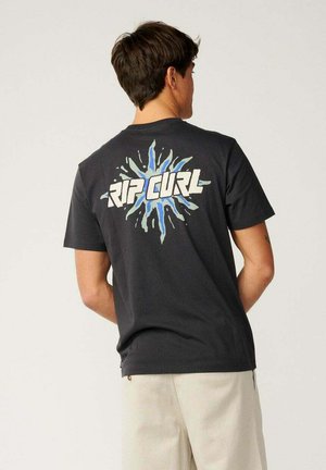 Młody mężczyzna zwrócony tyłem, ubrany w czarną koszulkę z niebiesko-białym logo "Rip Curl" oraz jasnobeżowe spodnie na jednolitym tle.