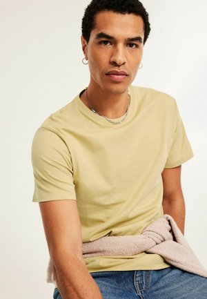 Junger Mann mit einem hellgelben T-Shirt, silberner Kettenhalskette, Creolenohrringen und einem beigefarbenen Pullover, der um seine Taille gebunden ist.