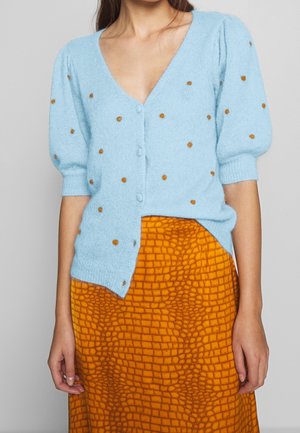 Žena ve světle modrém cardiganu s knoflíky, hnědými puntíky a oranžové texturované sukni s mřížkovým vzorem, stojící na šedém pozadí.