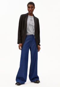 Vrouw in een zwart colbert, grijze overhemd met knopen, brede blauwe denim broek met trekkoord en zwarte fringeschoenen, staande tegen een witte achtergrond.