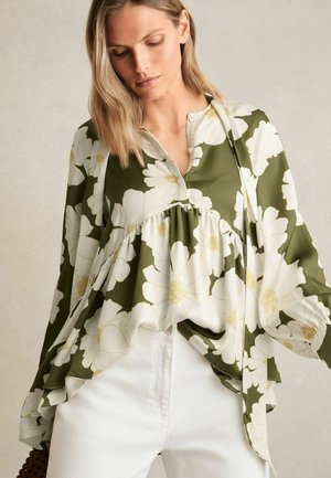 Groene bloemenblouse met witte bloemen, losse pasvorm, V-hals enGathered taille-details, gecombineerd met witte broeken. Gladde stofstructuur.