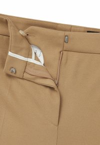Beige trousers with a smooth texture, featuring a zipper closure and button detail. Label visible inside with size information.