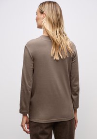 Pitkähihaisessa taupe-värisessä paidassa on yksinkertainen pyöreä kaula-aukko ja sileä pinta. Takana näkyy rento istuvuus ja hienovaraisia ompeluyksityiskohtia.
