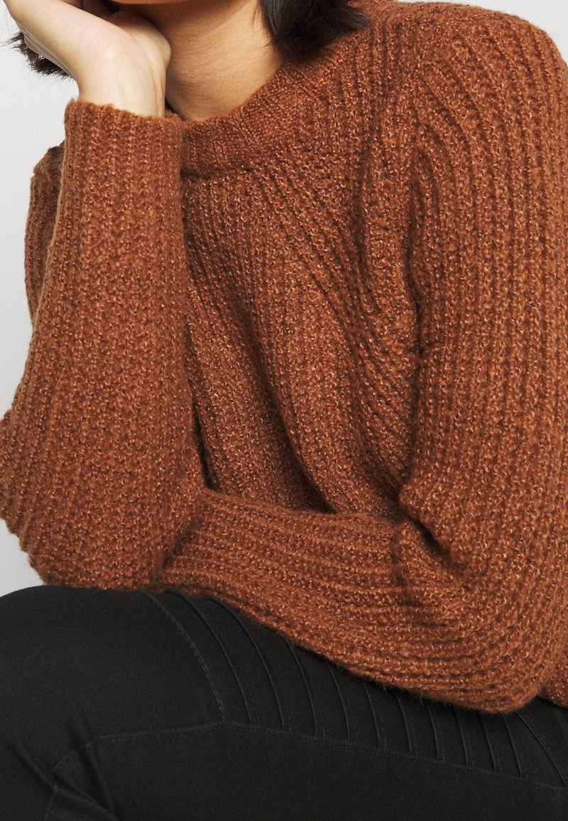 Femme portant un pull en maille marron texturé à manches longues, reposant son visage sur une main, assorti à un pantalon noir.