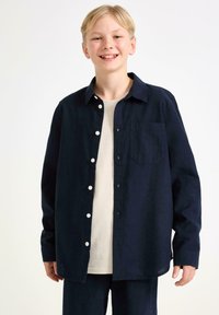 Camicia a bottoni blu navy con tasca, realizzata in un tessuto testurizzato. Presenta maniche lunghe e una vestibilità rilassata, indossata sopra una t-shirt di colore chiaro.