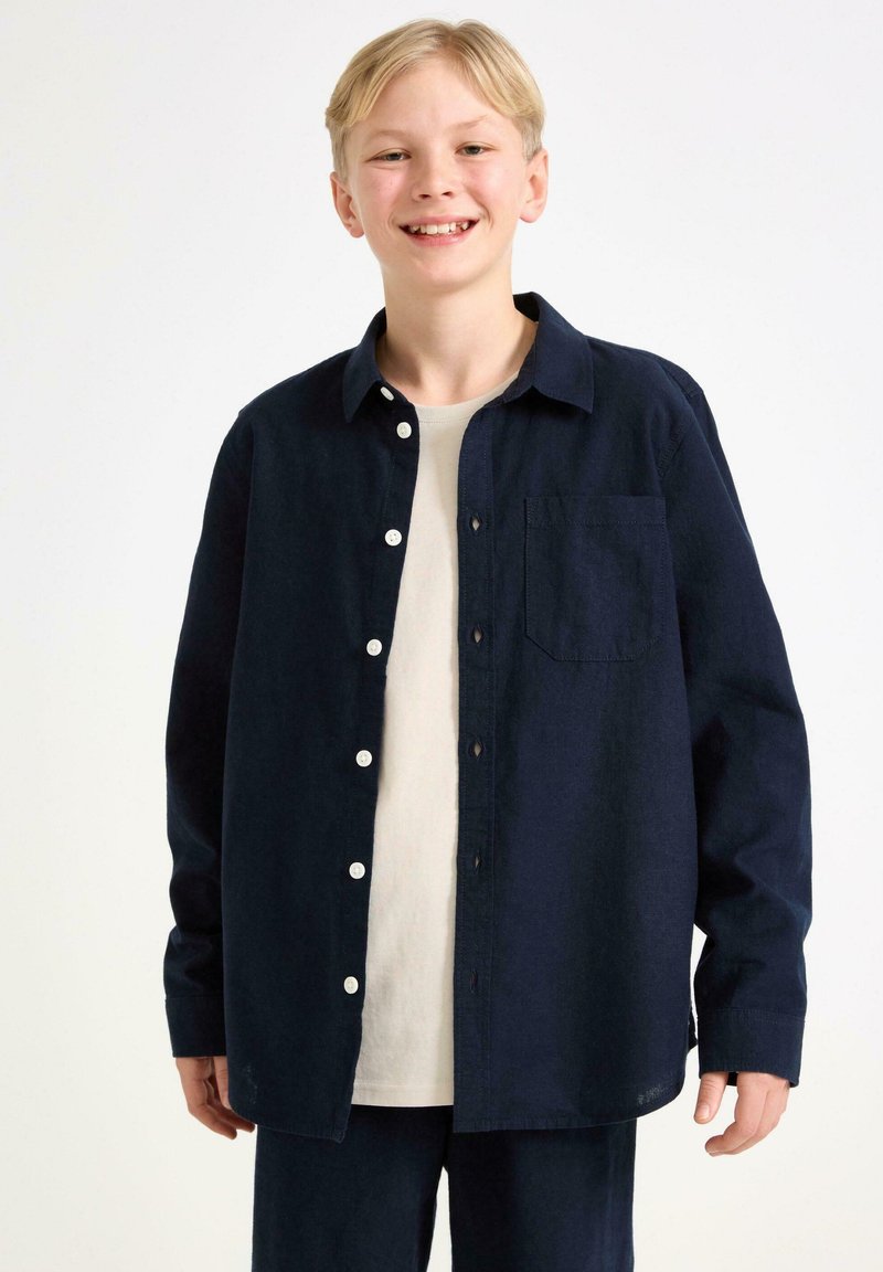 Camicia a bottoni blu navy con tasca, realizzata in un tessuto testurizzato. Presenta maniche lunghe e una vestibilità rilassata, indossata sopra una t-shirt di colore chiaro.