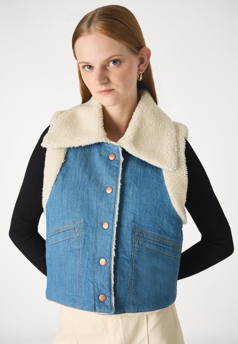 Kamizelka dżinsowa z kremowym kołnierzem z sztucznego owczego futra, pięcioma miedzianymi guzikami i dwiema przednimi kieszeniami. Charakteryzuje się niebieskim odcieniem z akcentami w postaci szwów.