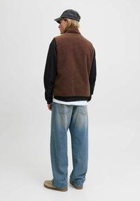 Person, der mit dem Rücken zur Kamera steht, trägt eine braune Fleeceweste, ein schwarzes Langarmshirt, lockere blaue Jeans, tanfarbene Schuhe und eine dunkle Baseballkappe.