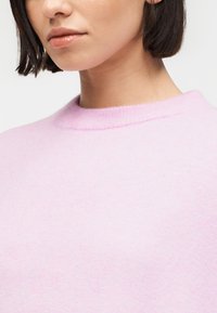 Γυναίκα με κοντά σκούρα μαλλιά, φορώντας ελαφρώς ροζ πουλόβερ με στρογγυλή λαιμόκοψη, τα χείλη ελαφρώς γυαλιστερά, μπροστά σε απλό φωτεινό φόντο.