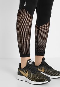 Nogi ubrane w czarne legginsy siateczkowe i czarne buty biegowe Nike z złotym logo na białej podłodze i jednolitym tle.