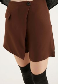 Pantaloni corti marroni con una texture liscia, dotati di chiusura a bottone e una tasca laterale. Il design include una pattina frontale e una vestibilità sartoriale.
