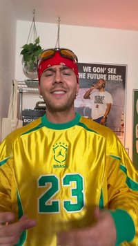 Homme portant un maillot de sport jaune du Brésil avec le numéro 23, un bandana rouge, des lunettes de soleil sur la tête, souriant à l’intérieur avec des affiches et une plante suspendue derrière.
