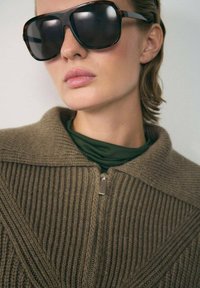 Gafas de sol negras con un grueso marco de carey, usadas con un suéter marrón de canalé con cremallera sobre una blusa verde. Textura suave y mate.