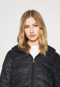 Čierna páperová bunda so zipsom a kapucňou, s prešívaným dizajnom a hladkým povrchom. Nosená nad bielym pleteným svetrom.