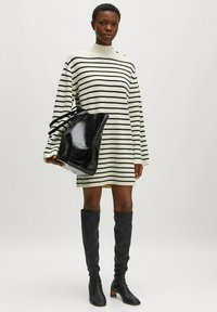 Robe pull en tricot avec des rayures noir et blanc, col montant, manches larges ; associée à un sac tote en cuir noir et des bottes noires jusqu'aux genoux.