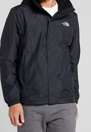 Mann trägt eine schwarze The North Face Jacke mit Kapuze, weißes Innenhemd sichtbar, und graue Hose vor einfarbigem Hintergrund.
