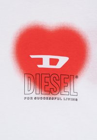 Balta T-kreklā ir sarkanas krāsas gradienta sirds dizains ar vārdu "DIESEL" melnā burtu izpildījumā un apakšā teksts "VEIKSMĪGAI DZĪVOŠANAI".