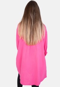 Übergroßes, hellrosa Shirt aus strukturiertem Stoff mit langen Ärmeln und abgerundetem Saum, getragen über schwarzen Hosen.