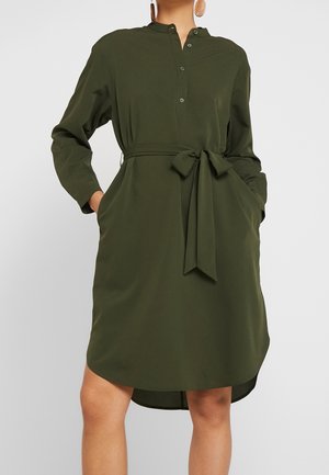 Vrouw draagt een knielange donker groene jurk met lange mouwen, knopen, zijzakken en een stoffen ceintuur geknoopt in de taille.