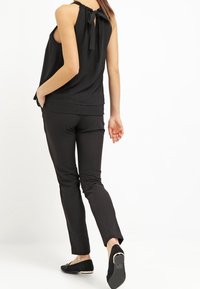 Czarny bezrękawnik z kokardą z tyłu, w połączeniu z wąskimi czarnymi spodniami i płaskimi butami. Gładka faktura, prosty design i jednokolorowa kolorystyka.