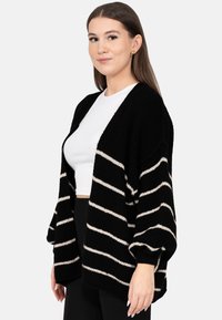 Młoda kobieta z długimi brązowymi włosami, nosząca złote kolczyki, biały krótki top oraz czarny kardigan z białymi poziomymi paskami, stojąca bokiem.