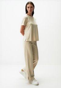 Beżowy t-shirt z krótkim rękawem i bocznymi rozcięciami, zestawiony z pasującymi spodniami w szerokim kroju, wykonanymi z miękkiego materiału. Całość dopełniają białe sneakersy.