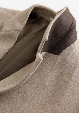 Beige kabát z vlněné směsi s tmavě hnědým límcem, s texturovaným vnějším povrchem a hladkým vnitřním podšívkou. Výrazné zakázkové klopy.