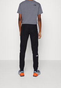 Grå kortärmad t-shirt och svarta träningsbyxor. Byxorna har en liten vit logotyp. Skorna är blå med orange detaljer och ett meshdizajn.