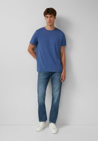 Blaues Rundhals-T-Shirt aus weichem Stoff, kombiniert mit hellblauen Jeans. Das Modell trägt weiße Sneaker und steht vor einem neutralen Hintergrund.