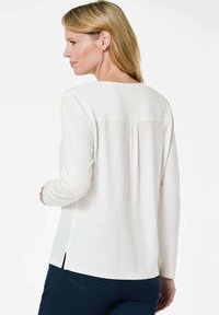 Weiße Bluse mit langen Ärmeln und rundem Halsausschnitt, ausgestattet mit einer Mittelrückenfalte und seitlichen Schlitzen, kombiniert mit dunklen Hosen. Glatte Stoffstruktur.