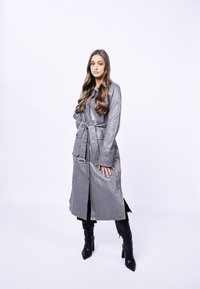 Lungo cappotto in pelle grigia con bottoni, una cintura e tasche; indossato sopra un vestito nero. La modella indossa stivali neri con tacco contro uno sfondo bianco.