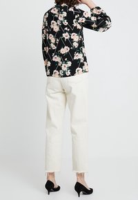 Blusa nera a fiori con fiori rosa e verdi, abbinata a jeans sfilacciati color avorio. Tacchi neri visibili in basso.