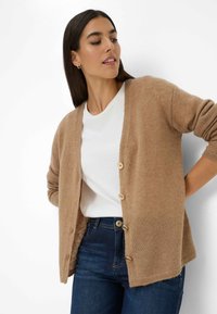 Cardigan marrone chiaro realizzato in morbido tessuto a maglia, con scollatura a V, chiusura a bottoni e polsini a costine, abbinato a una camicia bianca e jeans scuri.
