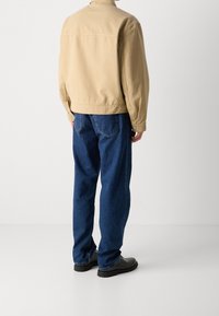 Beżowa kurtka z kołnierzykiem, zestawiona z luźnymi niebieskimi dżinsami. Kurtka ma guziki na rękawach i swobodny krój. Całość dopełniają czarne buty.