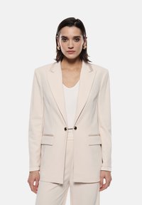Blazer beige chiaro con vestibilità su misura, due tasche frontali, bottoni con rifiniture dorate e cuciture a contrasto lungo il colletto e le lembi.