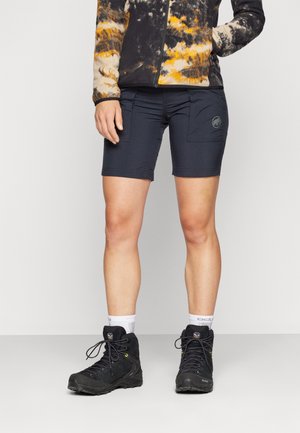 Person trägt schwarze Outdoor-Shorts mit Taschen, schwarze Wanderschuhe, weiße Socken und eine schwarz-orange gemusterte Jacke.