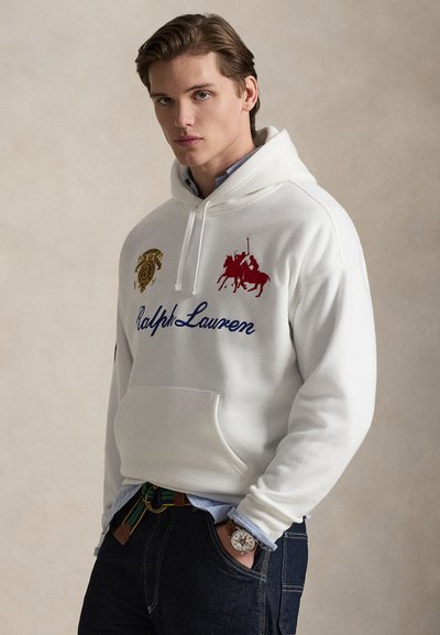 Jeune homme portant un sweat à capuche blanc Ralph Lauren avec logos, un jean foncé, une ceinture verte à rayures et une montre-bracelet, posant devant un fond uni.
