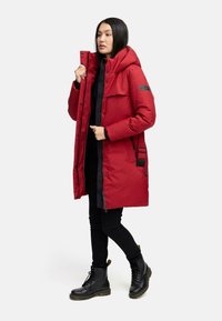 Red puffer coat with a high collar, zippered pockets, and a durable exterior. Features a front button closure and elastic cuffs.