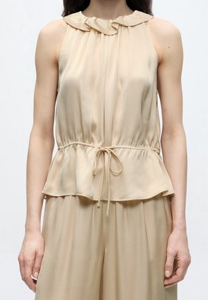 Femme portant un chemisier sans manches beige clair avec un col volanté et un lien à la taille, assorti d’un pantalon ample.