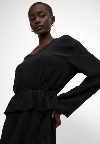 Musta mekko, jossa on V-pääntie, pitkät hihat ja nauhavyötärö. Valmistettu sileästä kankaasta, jossa on kerroksellinen peplum-yksityiskohta vyötäröllä.