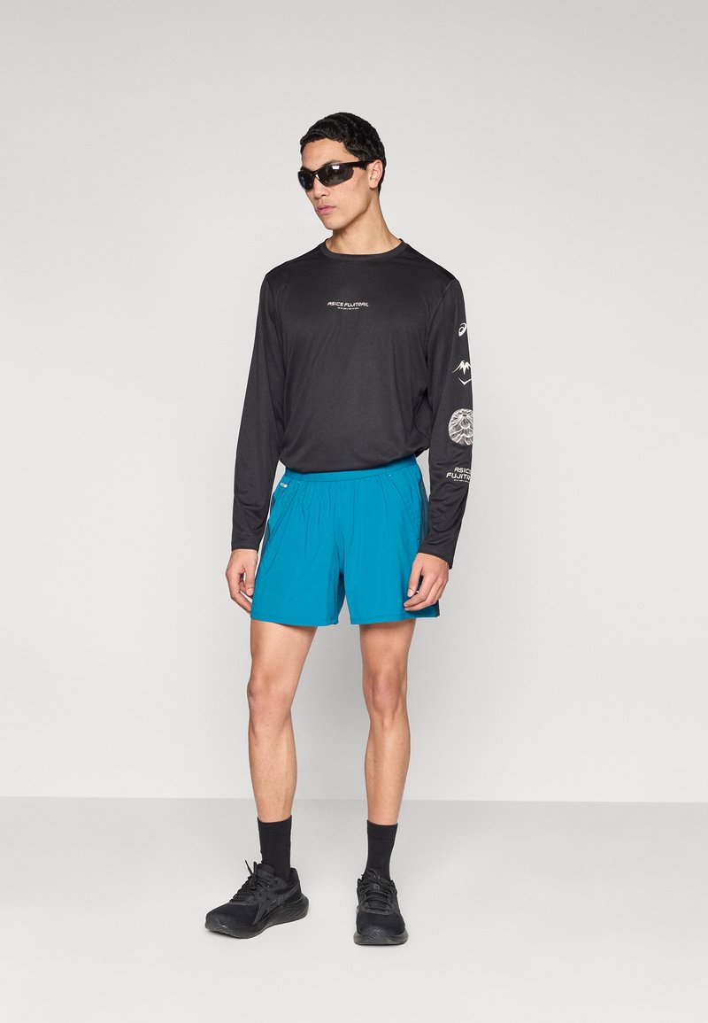 Chemise noire à manches longues avec impression blanche, short turquoise, chaussures noires et lunettes de soleil, présentant une coupe décontractée et une texture de tissu lisse.