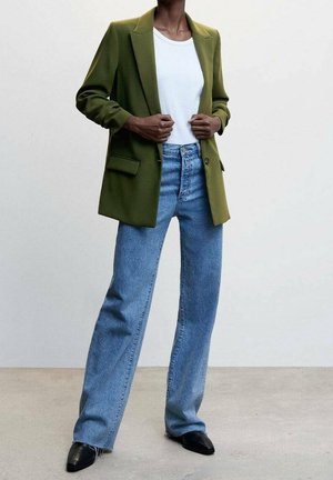 Person trägt grünen Blazer über weißem Top, blaue Jeans mit hoher Taille und schwarze spitze Schuhe und steht vor einem schlichten Hintergrund.