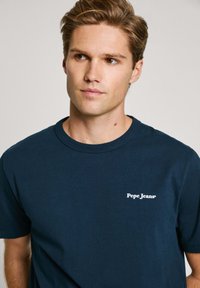 Tamno plava pamučna majica s okruglim izrezom i kratkim rukavima, s bijelim logom "Pepe Jeans" na prednjoj strani. Glatka tekstura.