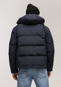 Granatowa kurtka puchowa z wysokim kołnierzem i odpinanym kapturem, o pikowanej strukturze i bocznych zamkach błyskawicznych. Nosi na niebieskich dżinsach.