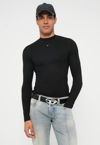 Schwarzes, langärmeliges, eng anliegendes Oberteil mit gerippter Textur und kleinem Logo. Kombiniert mit hellblauen Jeans und einem schwarzen Gürtel mit metallischer Schnalle.