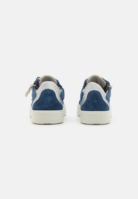 Baskets en suède bleue avec des accents blancs, une semelle plate et un détail en zip au niveau du talon. La vue de dos met en évidence la texture lisse et les coutures.