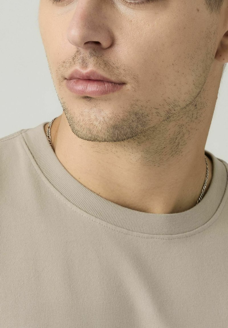 Primo piano della parte inferiore del viso di un uomo con una leggera barba, che indossa una maglietta beige con scollo rotondo e una collana di catena d'argento.