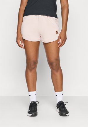 Roze sportshorts met elastische tailleband, gladde textuur en een klein logo-label. Draagt met zwarte sneakers en witte sokken.