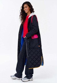 Žena s dlouhými kadeřavými vlasy se usmívá, má na sobě červený svetr, černý prošívaný dlouhý kabát s růžovou kapsou, tmavé džíny a bílé tenisky.