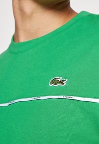Groene katoenen T-shirt met een ronde hals en korte mouwen. Bevat een witte horizontale band met de tekst "LACOSTE" en een geborduurd krokodillogo.